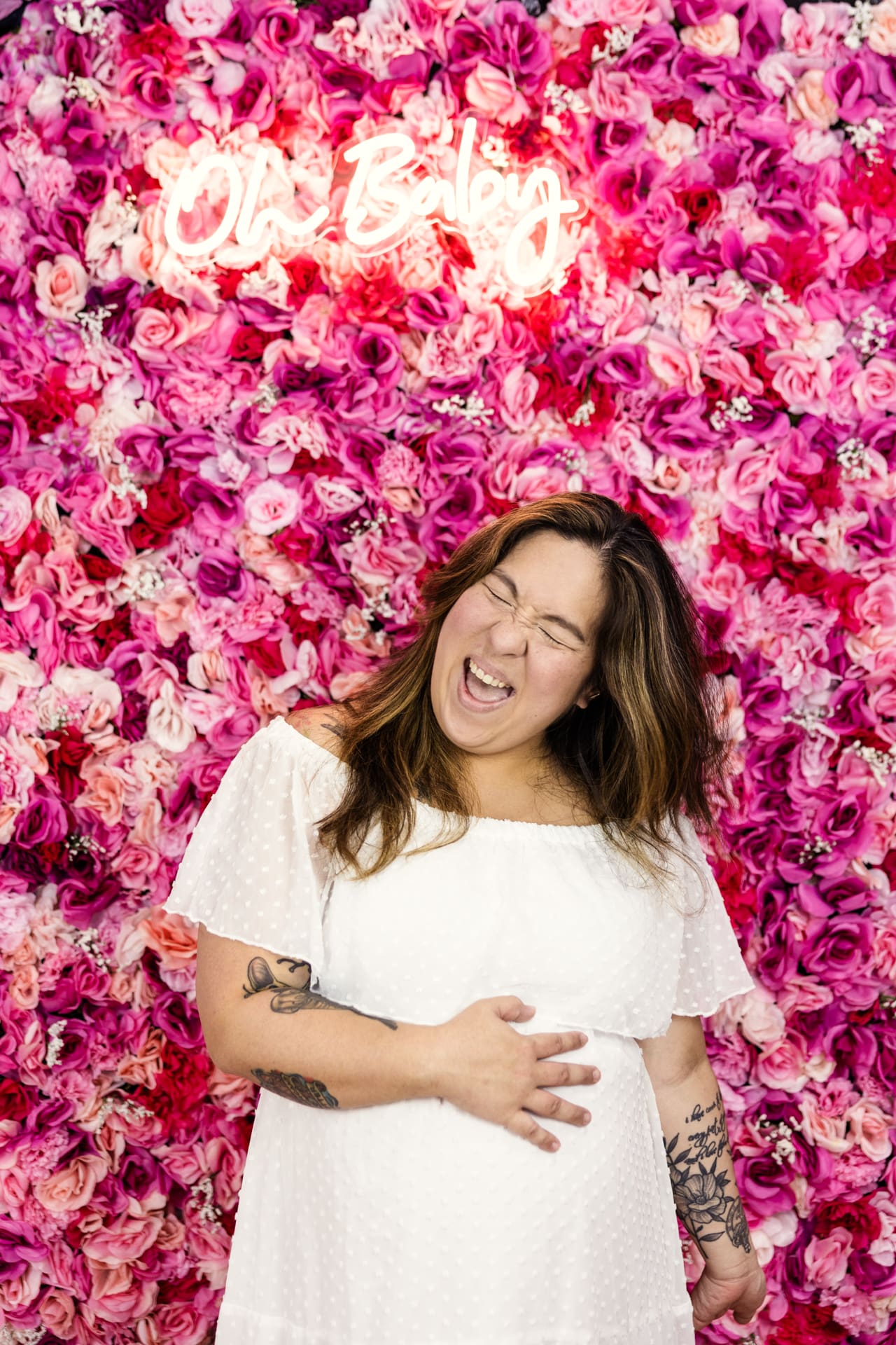 Candid photo of mom-to-be holding baby bump in front of pink flower wall with Oh Baby neon sign during maternity photoshoot at Chicago photography studio P&M Studio
