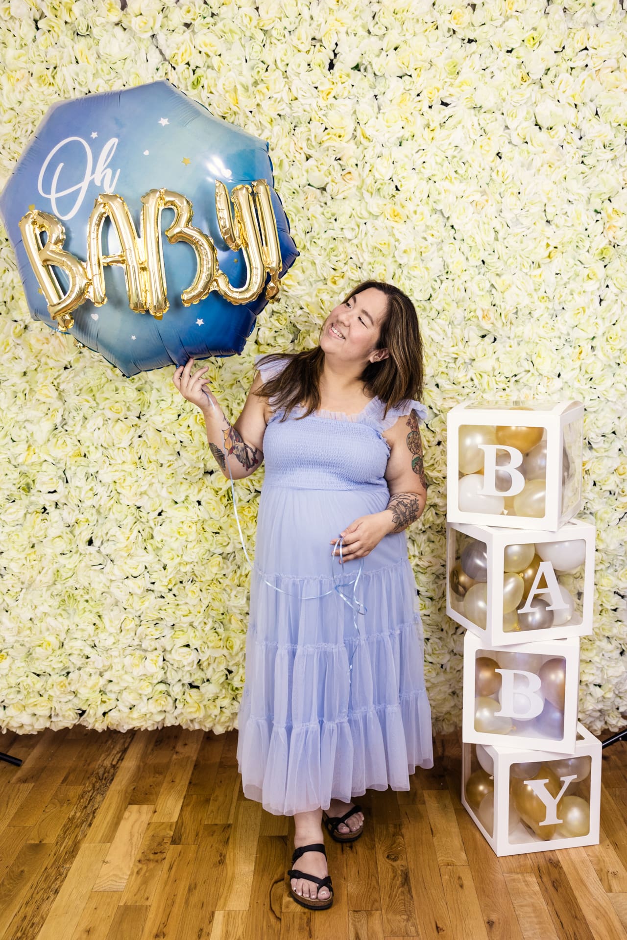 Candid photo of woman in blue dress holding Oh Baby balloon in front of floral wall during baby shower at Chicago private event space P&M Studio