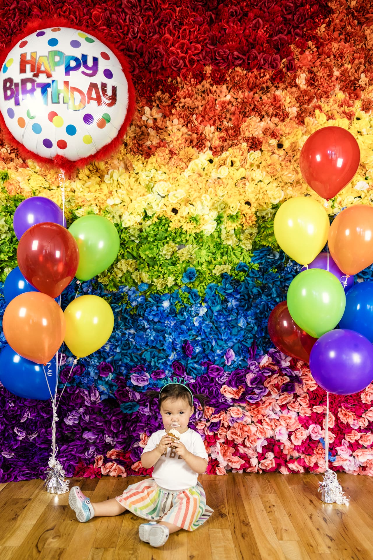Candid photo of toddler eating cupcake while sitting on floor in front of rainbow flower with Happy Birthday balloon at Chicago photography studio P&M Studio