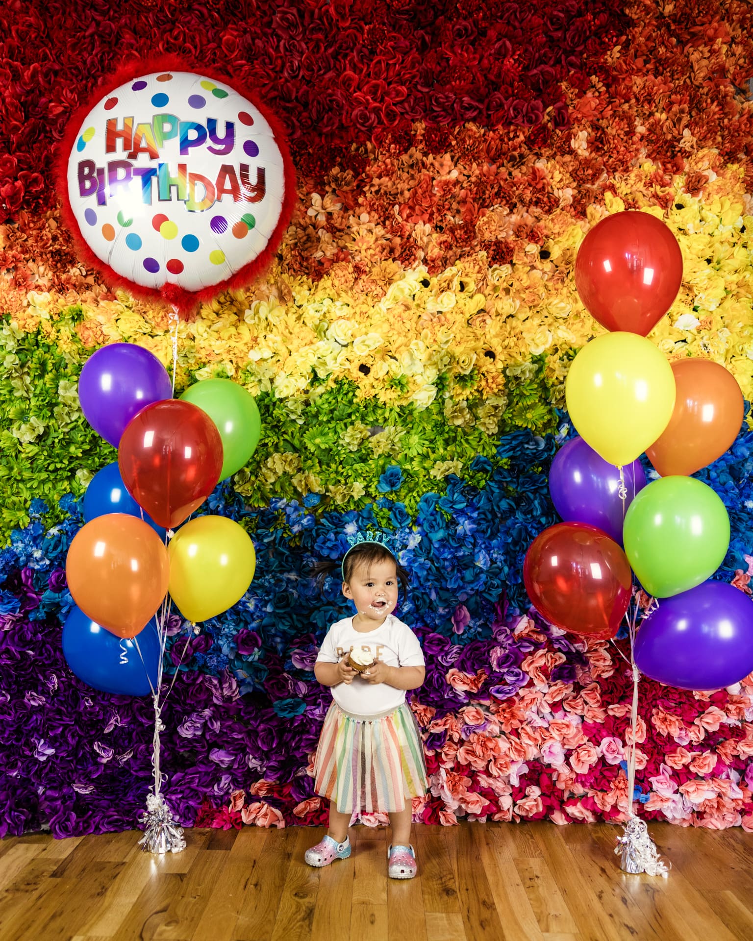 Toddler holds cupcake while standing in front of rainbow flower wall with multicolored balloons during birthday party photoshoot at Chicago photography studio P&M Studio