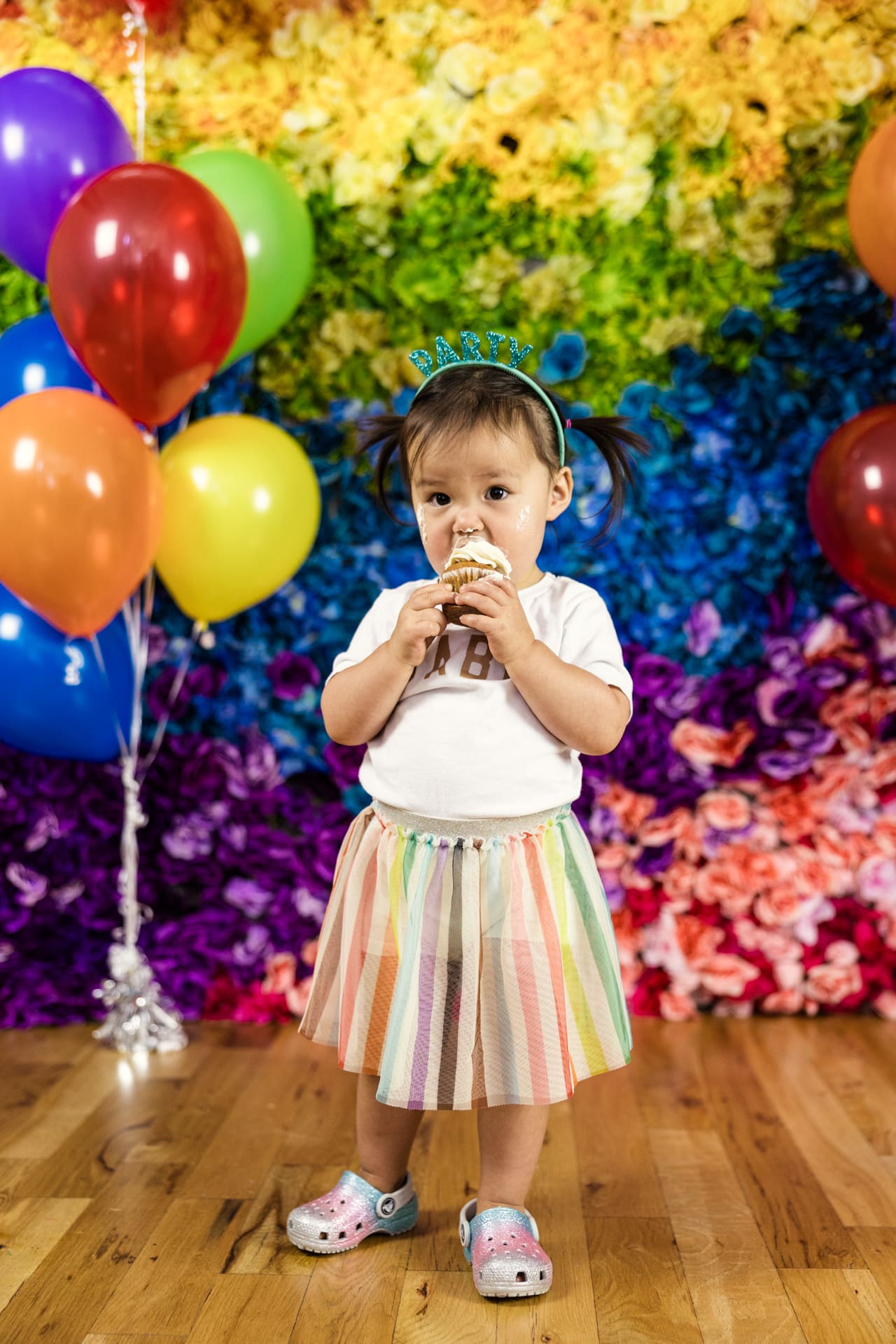 Shy toddler holds cupcake in front of rainbow flower wall with multicolored balloons during birthday party photoshoot at Chicago photography studio P&M Studio
