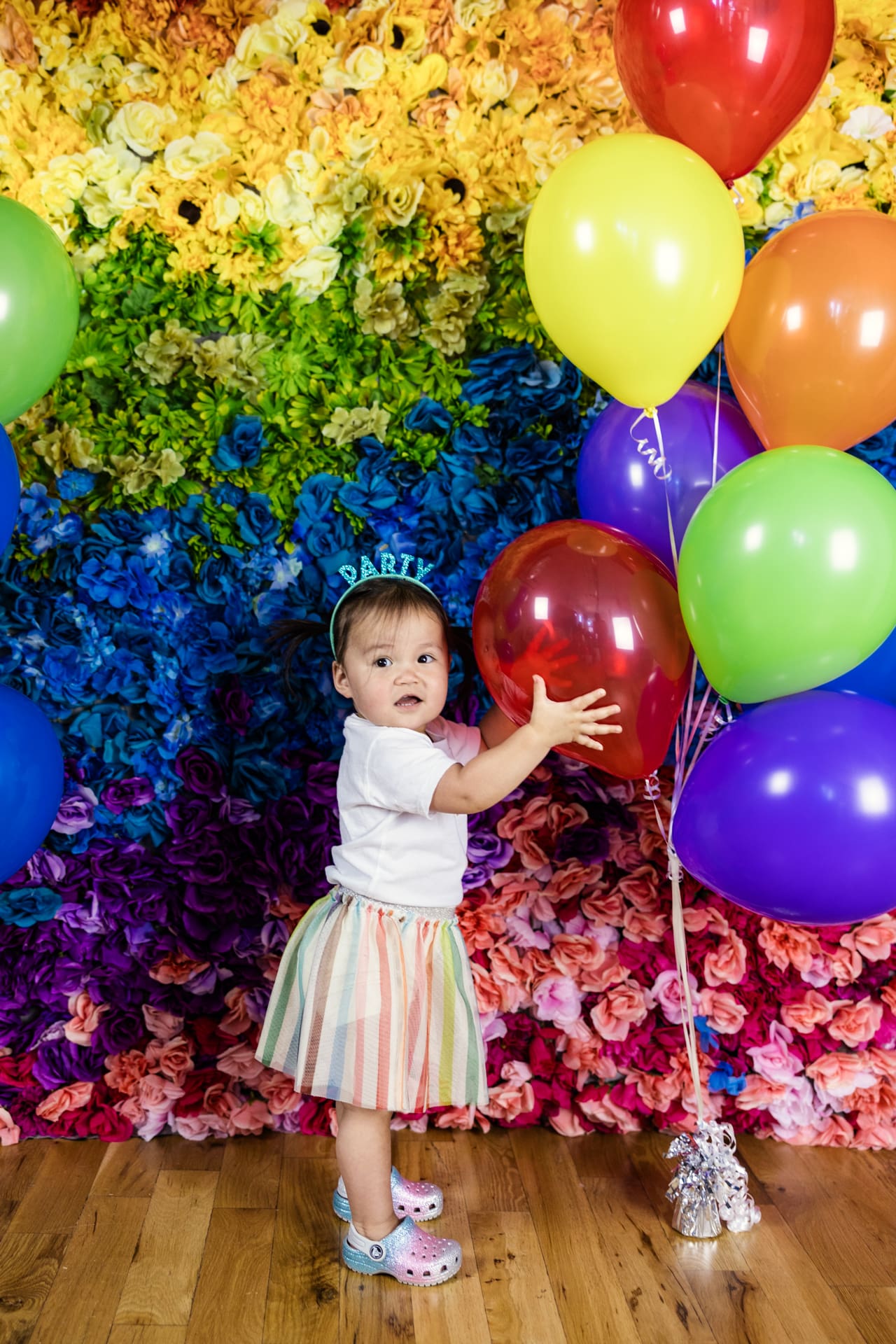 Candid photo of toddler holding red balloon in front of rainbow flower wall during birthday party photoshoot at Chicago photography studio P&M Studio