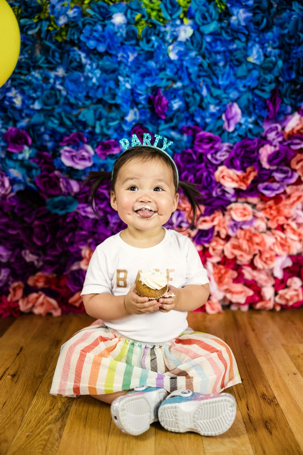 Happy toddler sits on floor holding cupcake in front of rainbow flower wall during birthday party photoshoot at Chicago photography studio P&M Studio