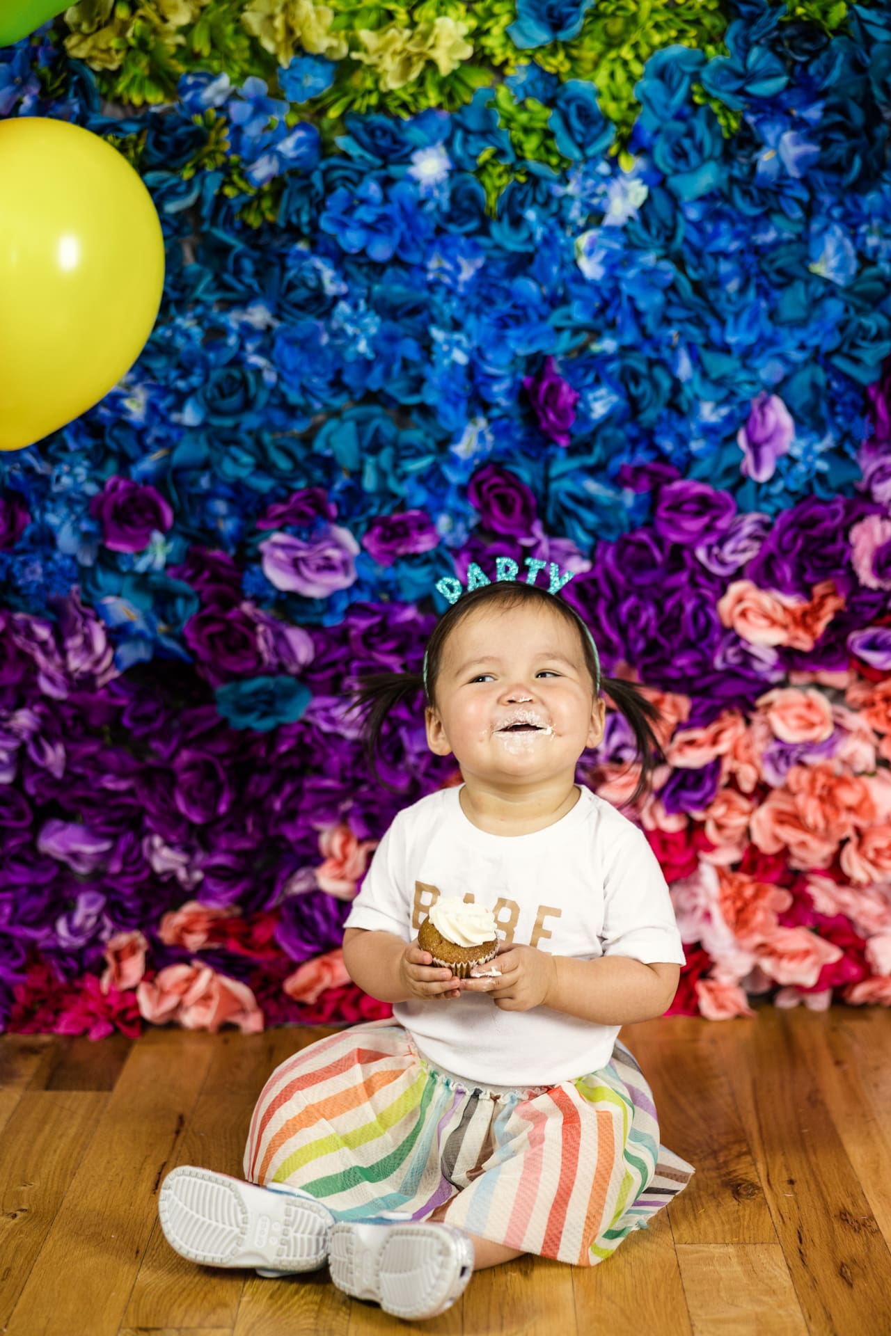 Candid photo of happy toddler eating cupcake in front of rainbow flower wall during birthday party photoshoot at Chicago photography studio P&M Studio