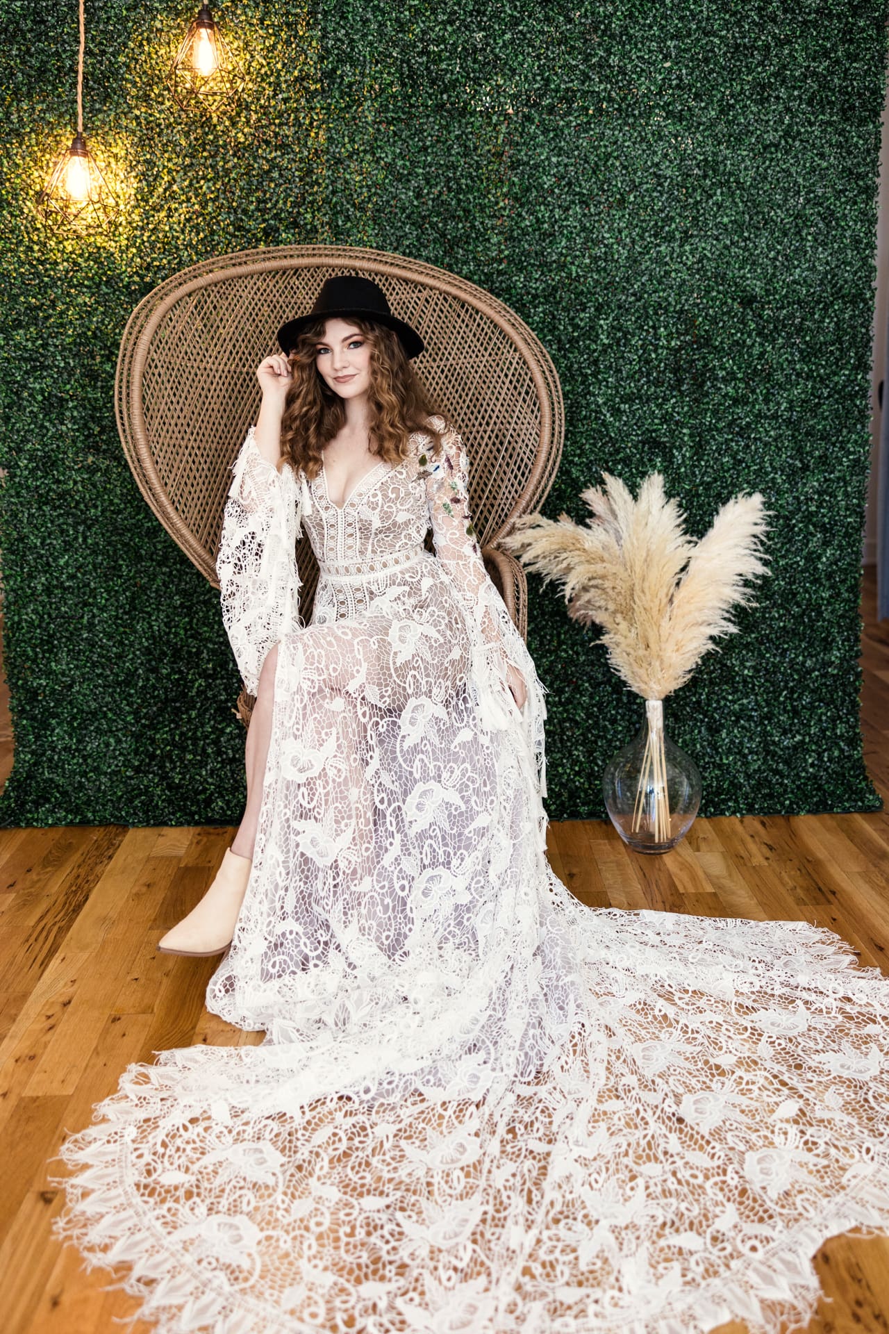 Model wearing long white lace dress and black fedora sits in wicker chair in front of greenery wall with pampas grass during fashion photoshoot at Chicago photography studio P&M Studio