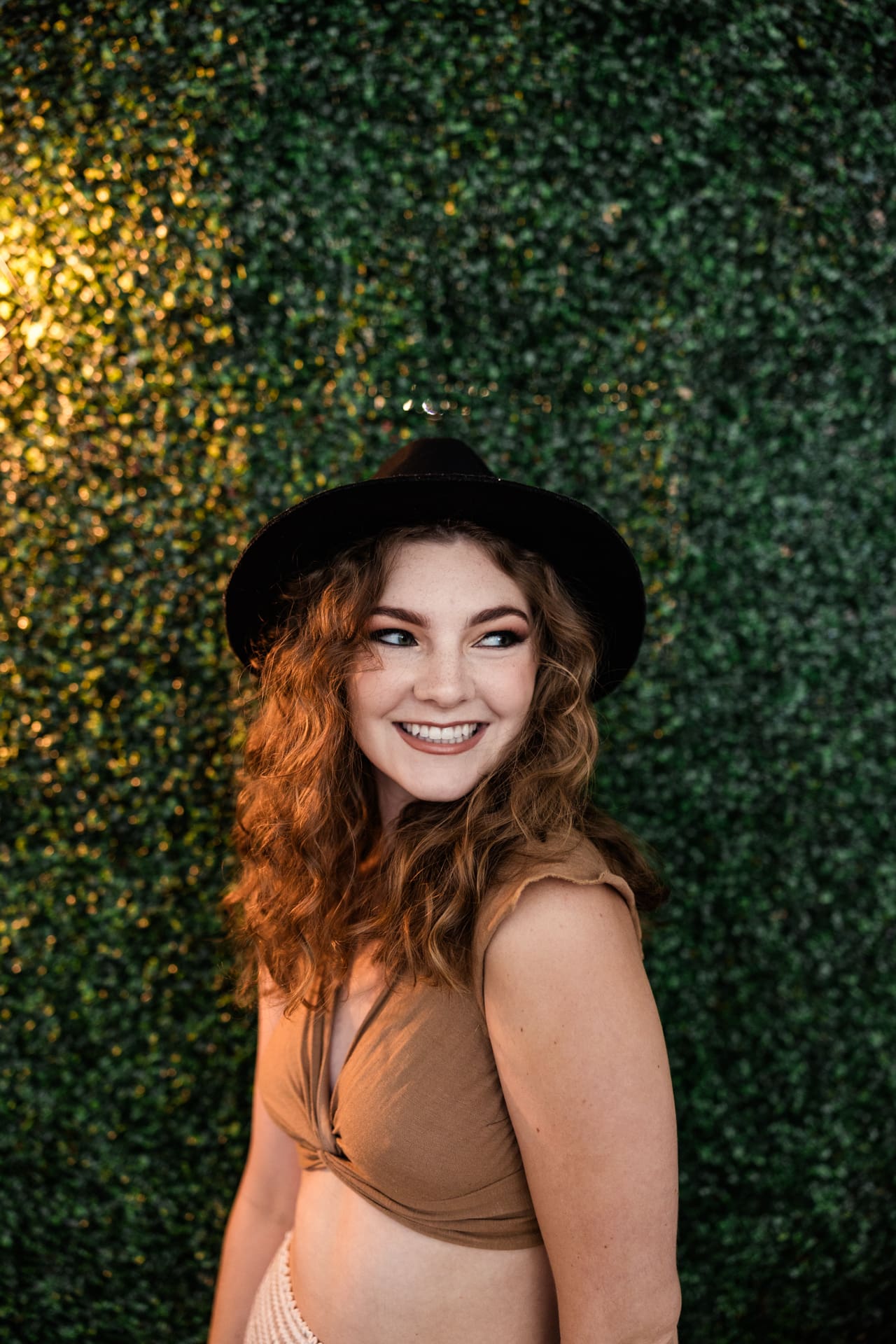 Candid photo of boho model wearing crop top and floppy black hat in front of greenery wall at Chicago photography studio P&M Studio