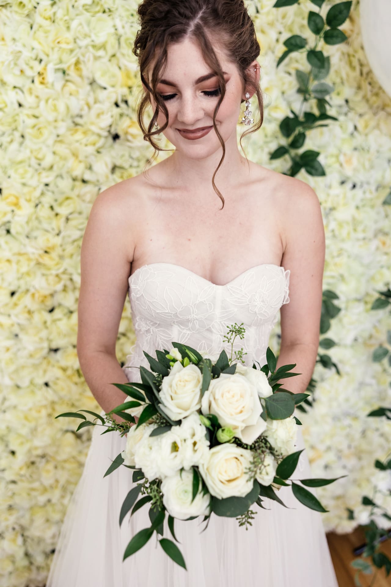 Classic photo of bride looking at white floral bouquet with greenery in front of white floral backdrop at Chicago photography studio P&M Studio