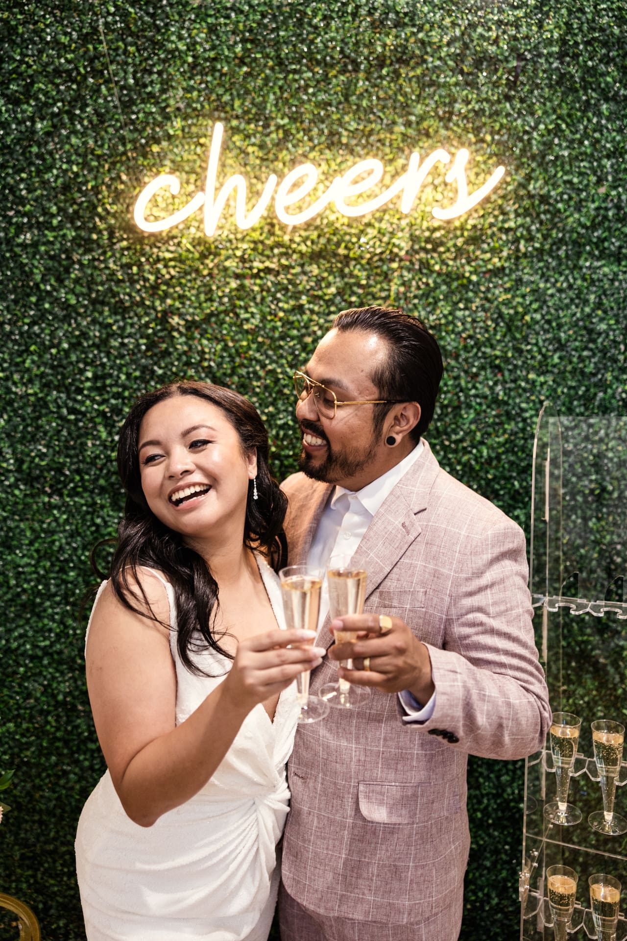Just married bride and groom toast champagne in front of greenery wall with Cheers neon sign after their intimate wedding ceremony at P&M Studio Chicago event space in West Town