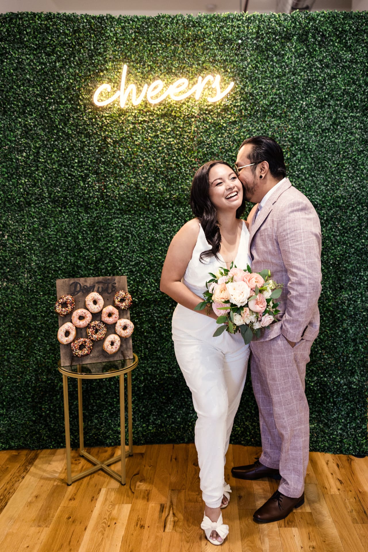 Just married bride and groom in front of greenery wall with Cheers neon sign and donut wall after their intimate wedding ceremony at P&M Studio Chicago event space in West Town