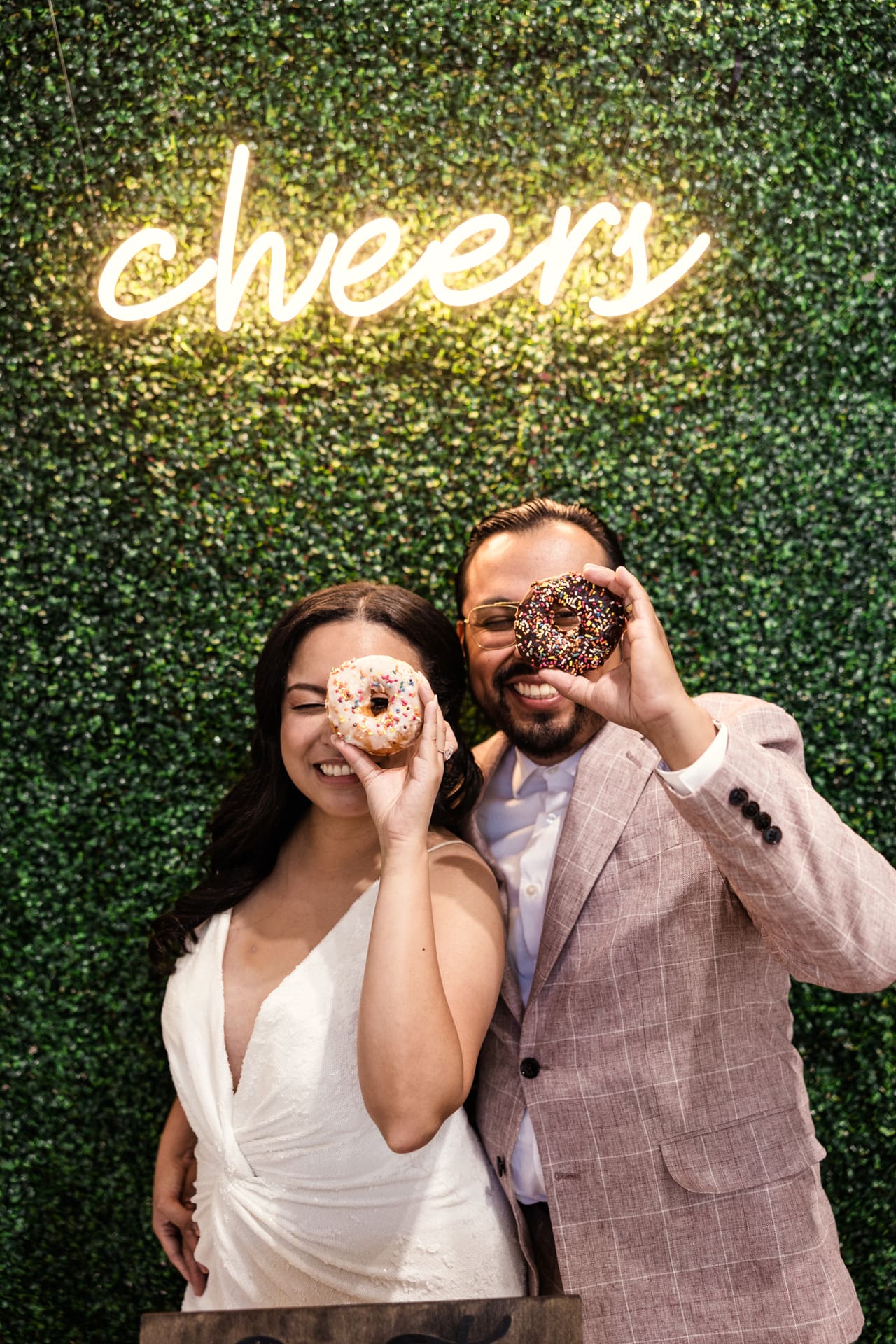 Fun photo of bride and groom holding donuts in front of greenery wall with Cheers neon sign after their intimate wedding ceremony at P&M Studio Chicago event space in West Town