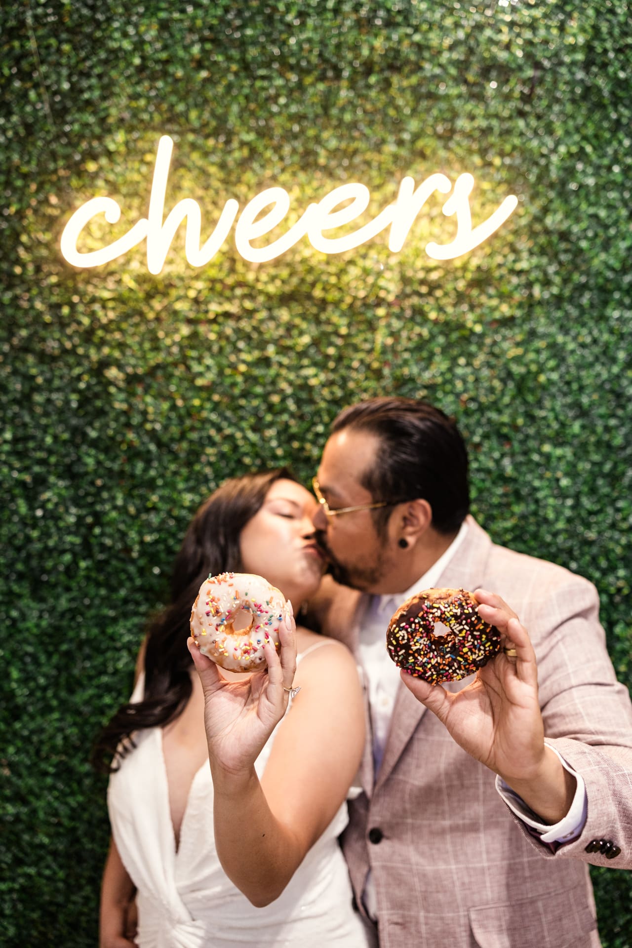 Fun photo of bride and groom holding donuts while kissing in front of greenery wall with Cheers neon sign after their intimate wedding ceremony at P&M Studio Chicago event space in West Town
