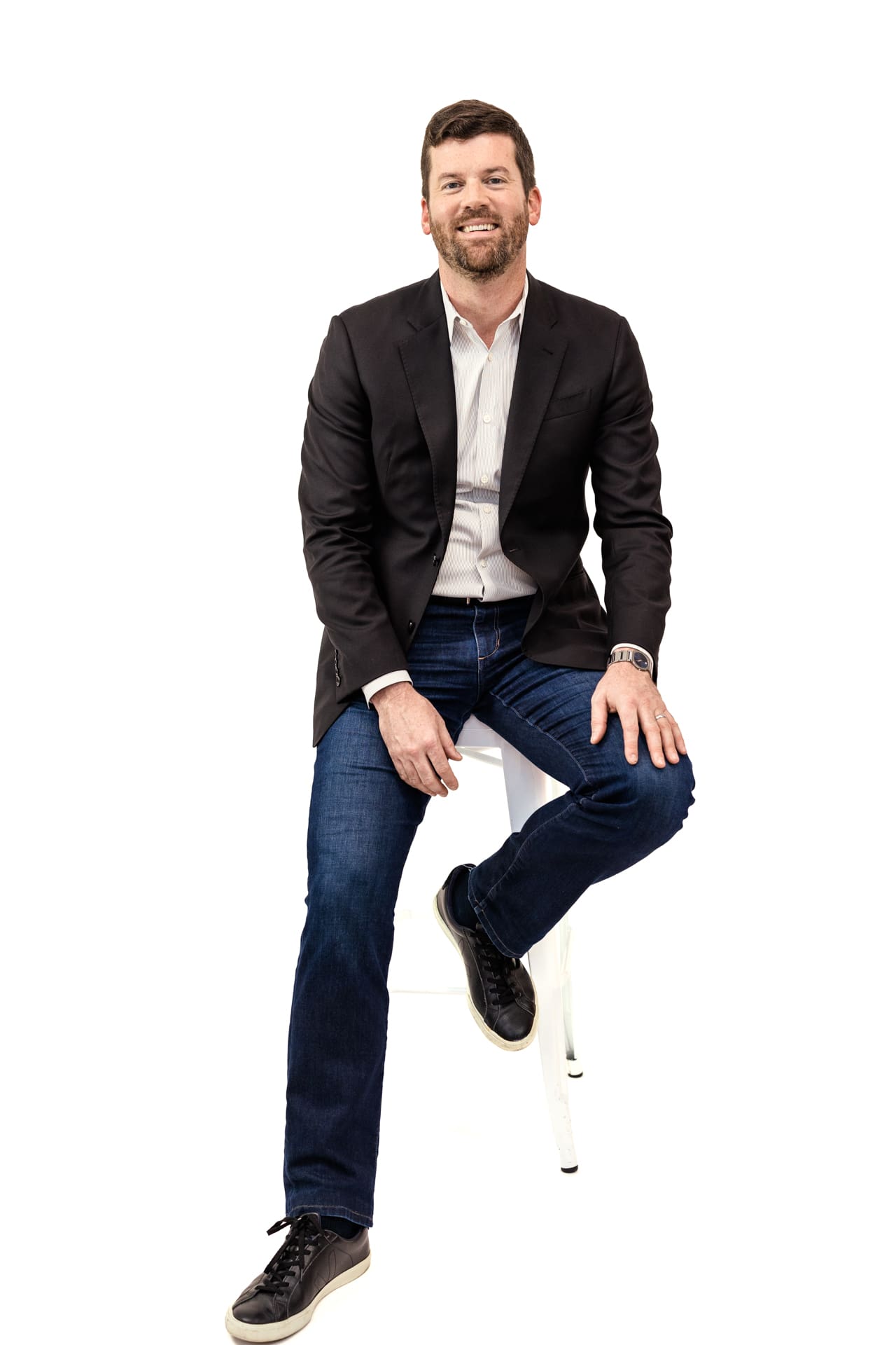 Studio portrait of man wearing suit jacket and dark denim pants with black sneakers on white backdrop at Chicago photography studio P&M Studio